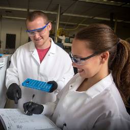 Male and female students in white lab coats and safety glasses, holding lab equipment and recording data in a logbook