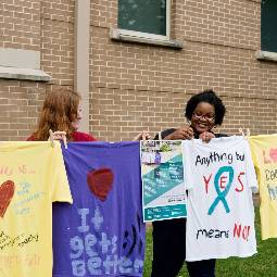 Two female students hang newly decorated T-shirts to dry on a clothesline outdoors, with messages in support of health-related issues