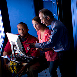 Professor pointing to computer screen, explaining something to a male and a female student who are gazing at the screen
