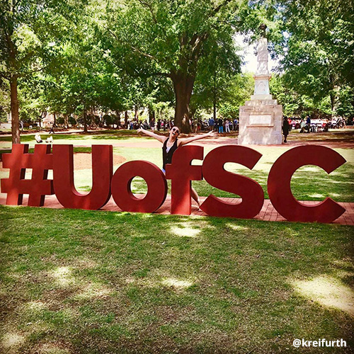Female student poses with a big smile and arms outstretched behind oversized garnet #UofSC letters on the Horseshoe.