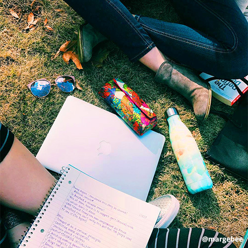 Two students studying with open notebook, laptop, water bottle, sunglasses laying on grass between them.
