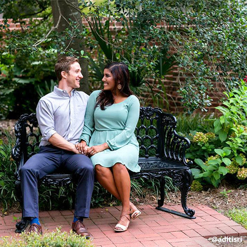 Young couple sits close to one another on bench in garden, looking at one another, smiling and holding hands.