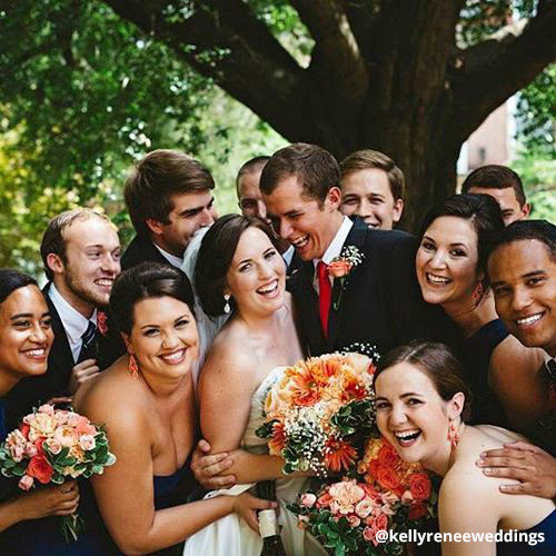 Wedding party surrounds smiling bride and groom, bride and bridesmaids hold colorful bouquets of flowers.