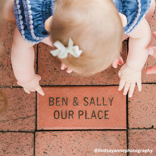 Toddler girl sits on brick sidewalk pointing to a brick that says, “Ben & Sally, Our Place.”