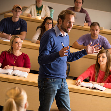 Professor lecturing in a classroom with students sitting in tiered rows of tables.