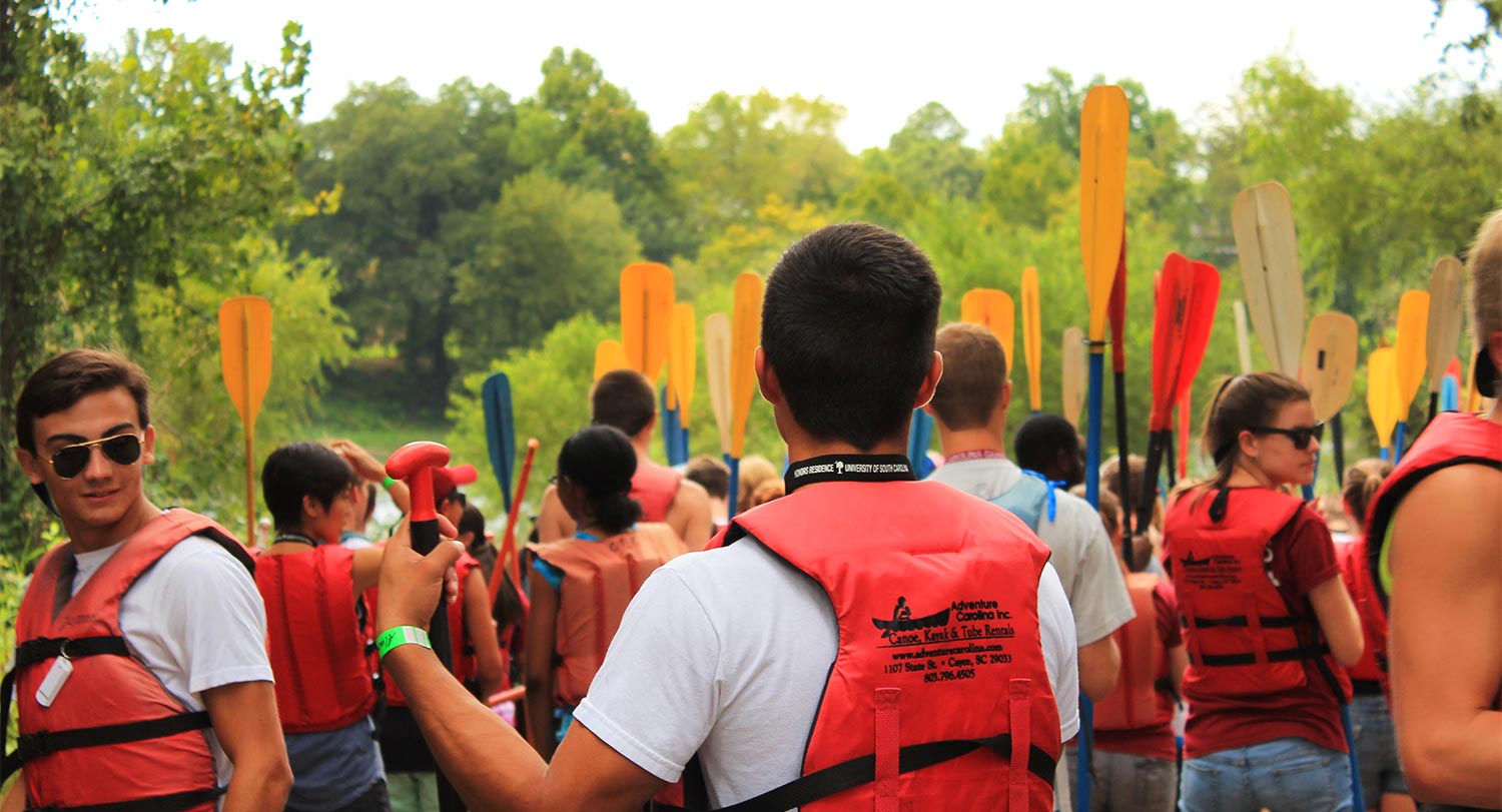 Students standing on the river bank with life vests and kayak paddles. 
