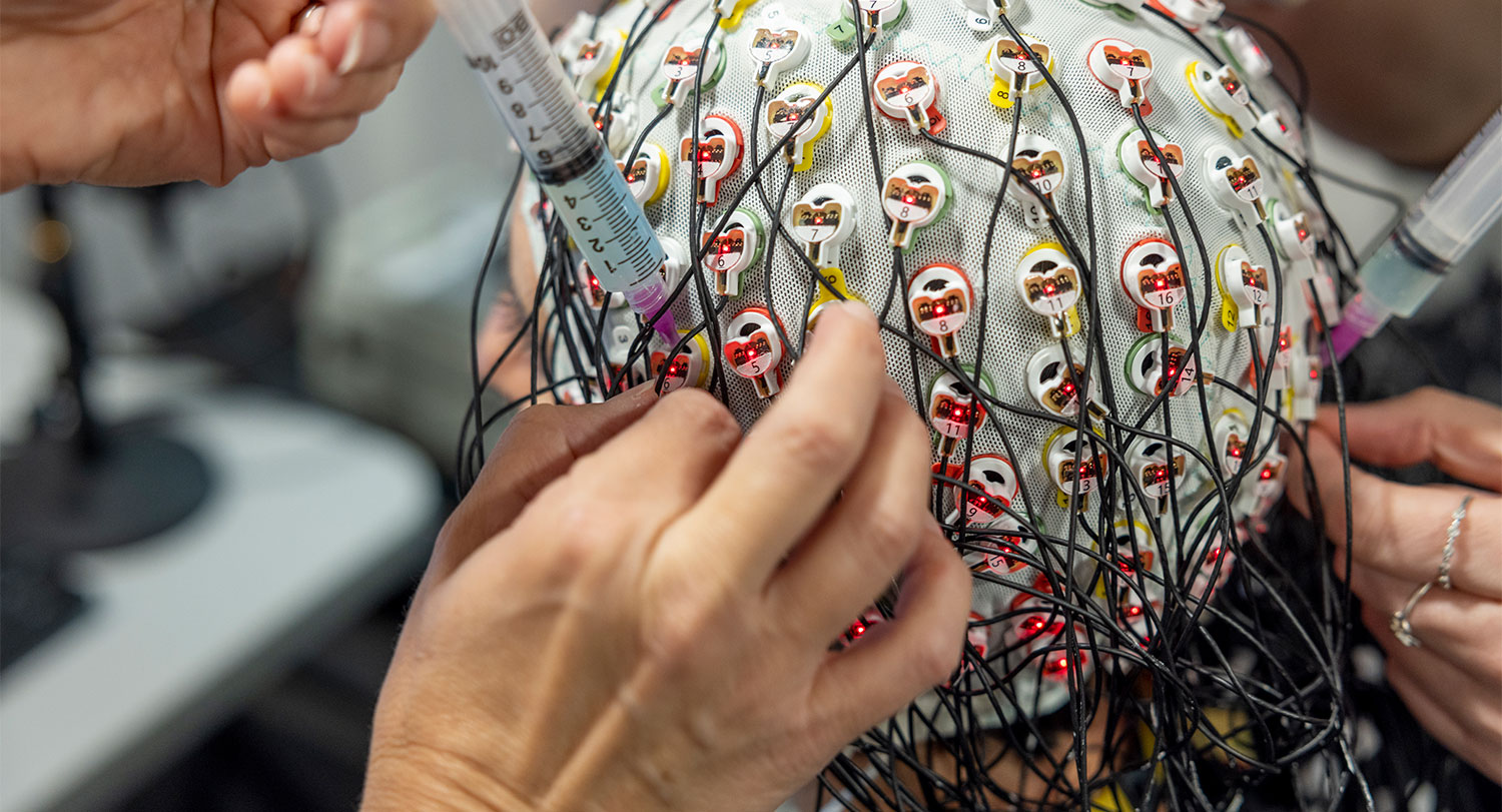 Hands adding a syringe of fluid into cap full of sockets and wires on a person's head.