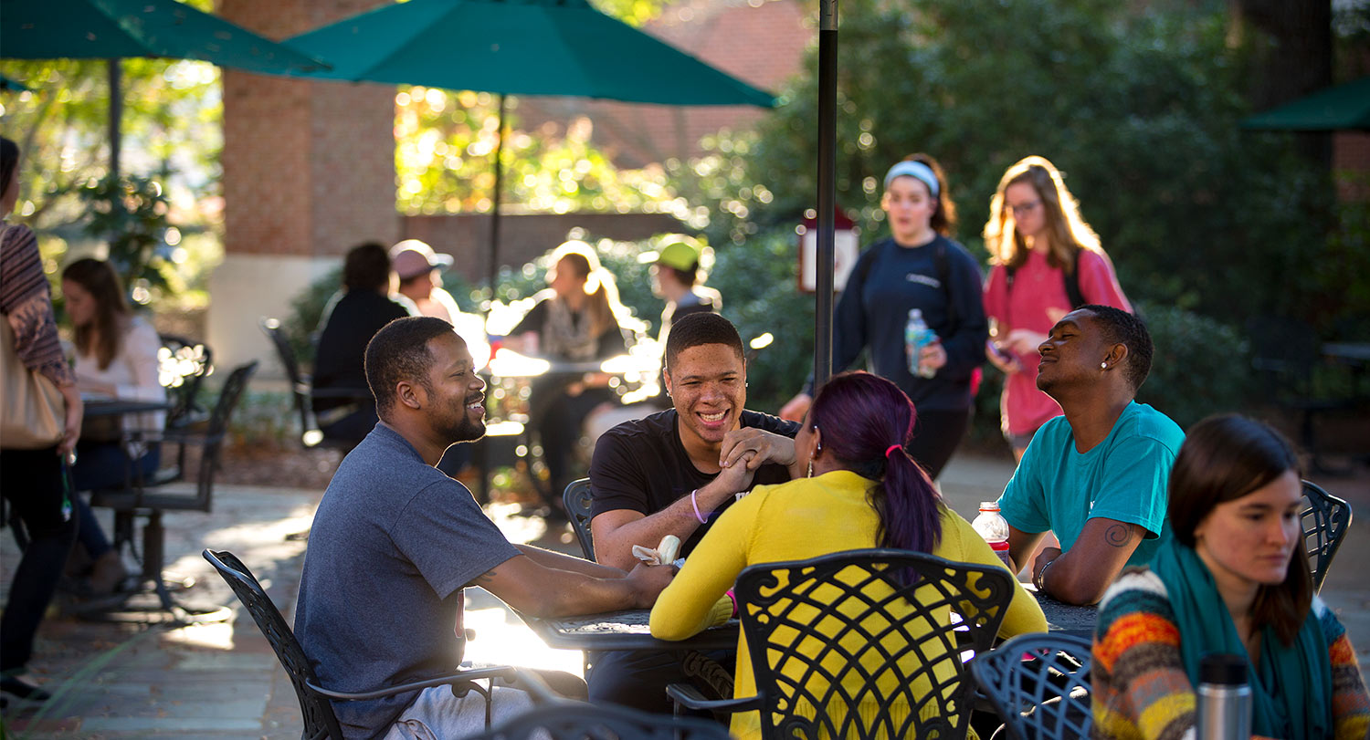 Group of students sitting at an outdoor patio table with other students mingling and walking around them.