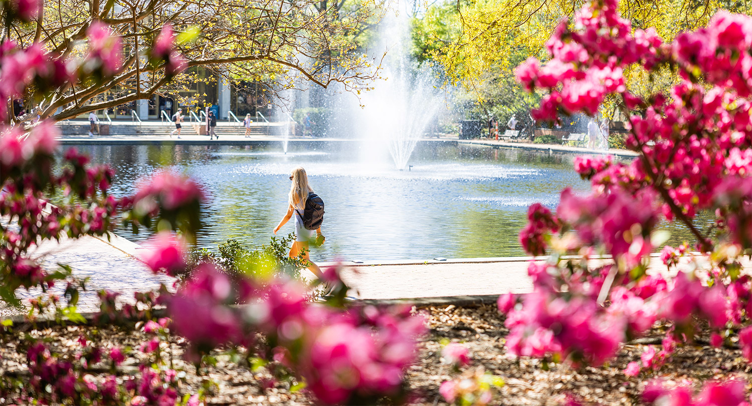 A student walking in front of a fountain running in the reflection pond with bright colorful flowers in the foreground. 