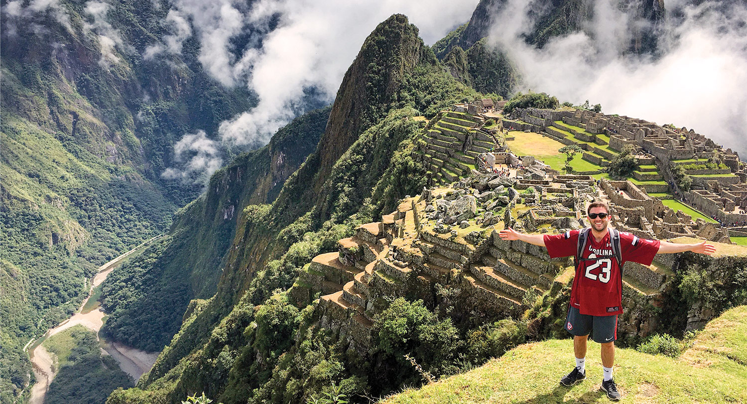 Student in a garnet South Carolina jersey standing in front of Machu Picchu with his arms stretched out.