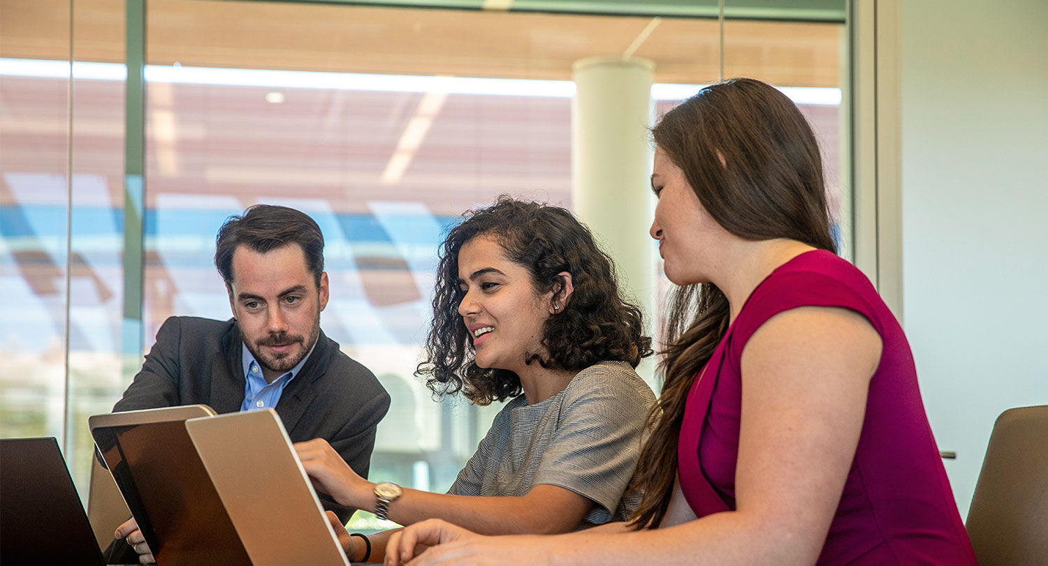 Three students working together at a conference room table with laptops.