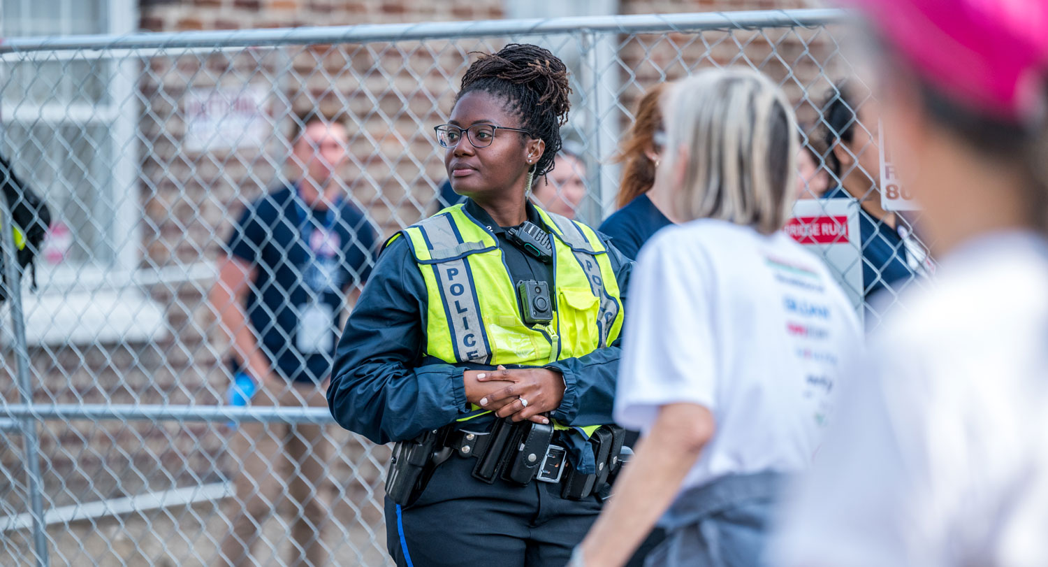 A police officer wearing a high-visibility vest and body camera stands in front of a chain-link fence, observing a public event.