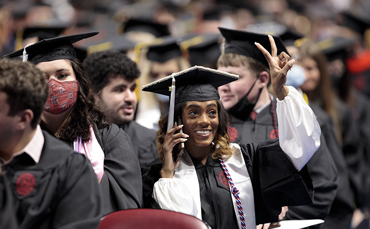 a young woman talking on a cell phone in a crowd of graduates holds up a peace sign to someone off in the distance