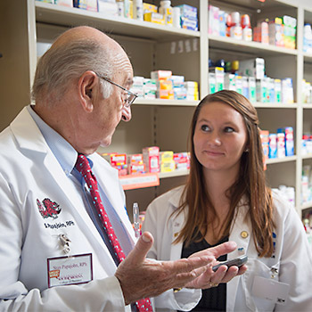 Student and a professor standing in front of a shelf of medication having a conversation.