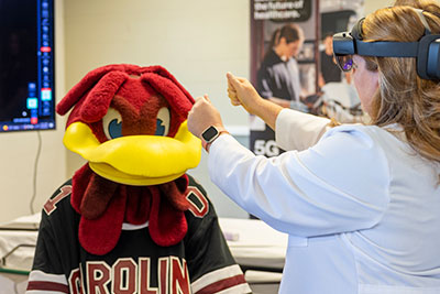 A person in a lab coat wearing virtual reality goggles holding her hands up in front of Cocky in a clinical setting.