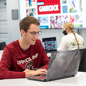 A student smiles while working on their computer as another student works in the background.