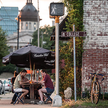 Group of people sitting at an outdoor dining table under a patio umbrell near a street sign that reads College Street and a view of the Statehouse in the background. 