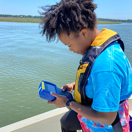Graduate student reading a water meter on a boat in a marsh.