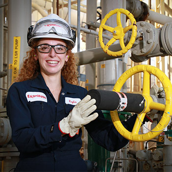 Student wearing a hard hat, safety goggles and Exxon Mobile coveralls on standing near equipment.