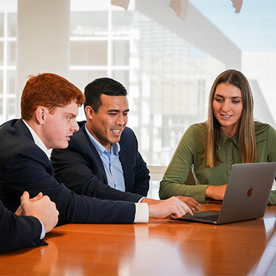 Three students in business clothes sitting around a laptop. 