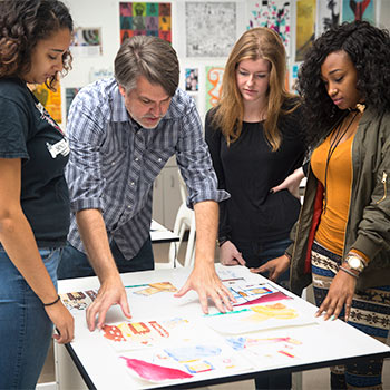 Students and a professor gathered around a table critiquing their artwork.