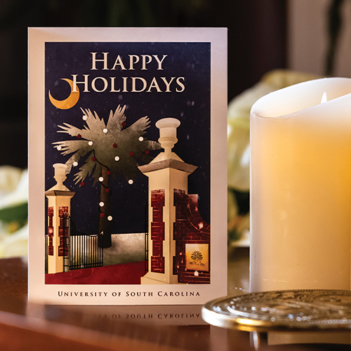 The USC 2023 holiday card, featuring an illustration of the Horseshoe gates, a palmetto tree and a crescent moon, sits on a table next to a white candle. 