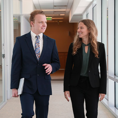 Two people in buisness attire walking down the hall talking.