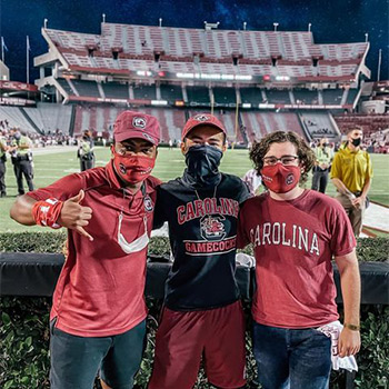 Three students in Gamecock gear and standing infront of the football field.