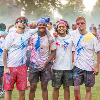 Four students covered in colored chalk dust from a Color Run smile with their arms around each other. 