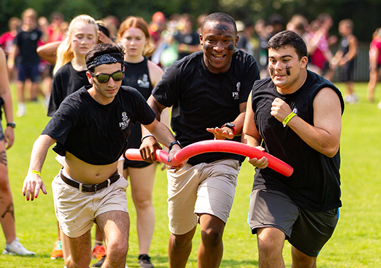 Three students running together in a relay race outside on a field.
