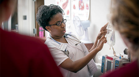 A nursing professor holding an IV bag instructing students in garnet scrubs. 