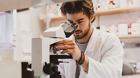 A student looking into a microscope wearing a lab coat. 