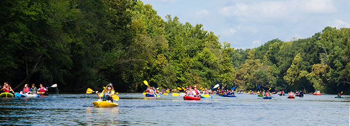 A river full of people paddling in colorful kayaks surrounded by lush trees on the river bank.