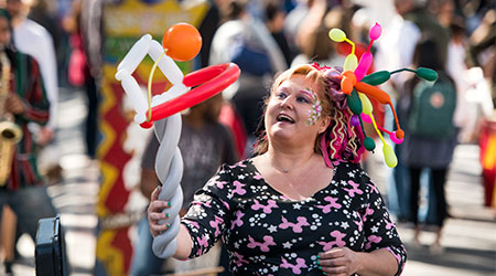A person holding a balloon animal wearing face paint at an outdoor market. 