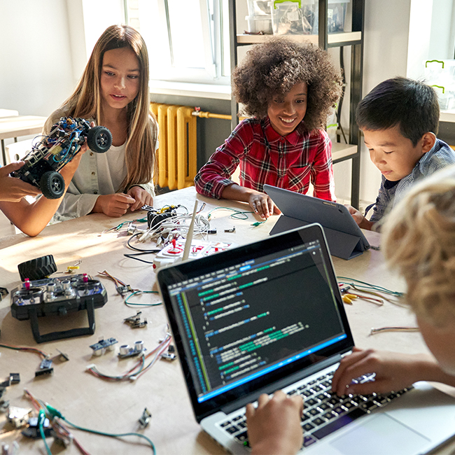 Group of children at a table putting together robotic cars and code on a laptop screen.