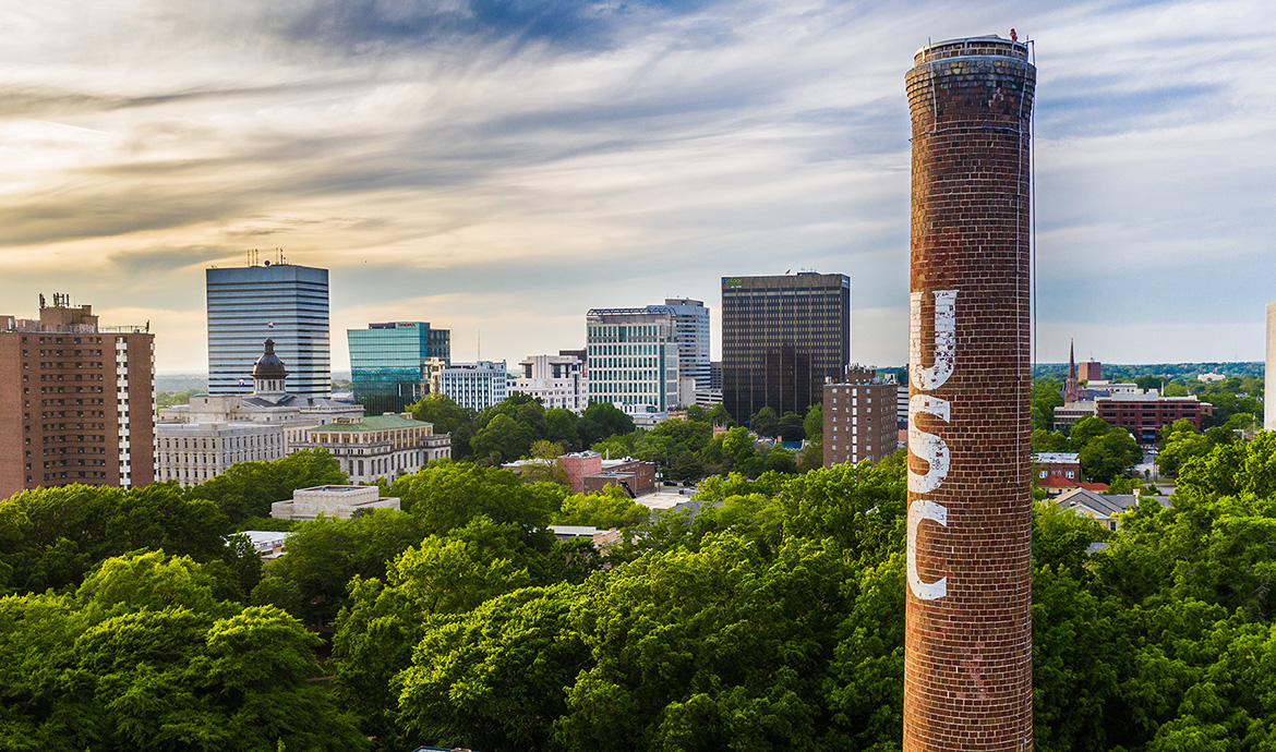 An aerial view of the University of South Carolina campus with tall buildings in the background and a brick smokestack with the letters USC printed vertically.