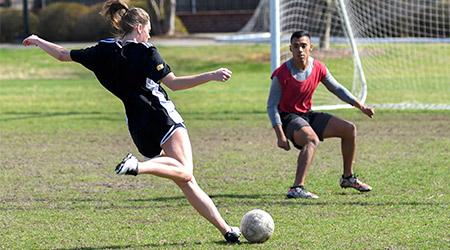 Two students playing on the co-ed soccer team, the player in the foreground is kicking the ball while the other defends the goal.