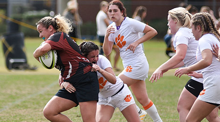 A women's intramural rugby player holds the ball while Clemson player tries to tackle her. 