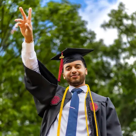 Graduate with his arm raised waving his fingers at the camera. 