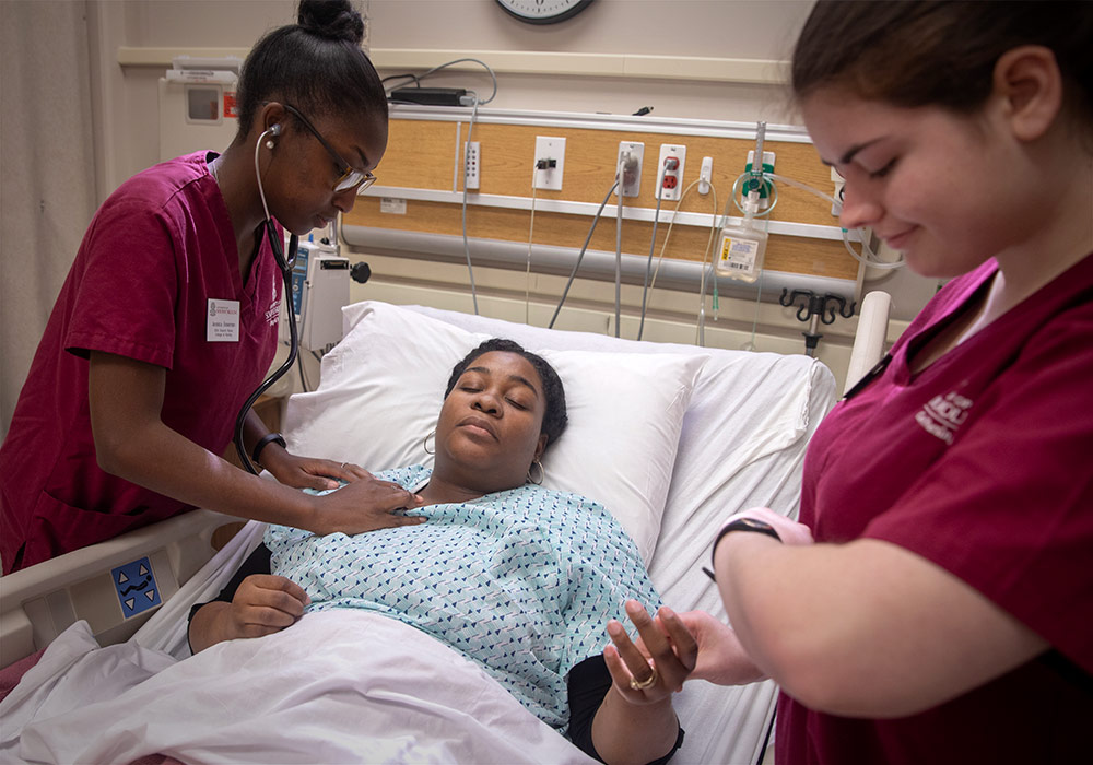 Two nursing students lean over a hospital bed taking a patients vitals. 