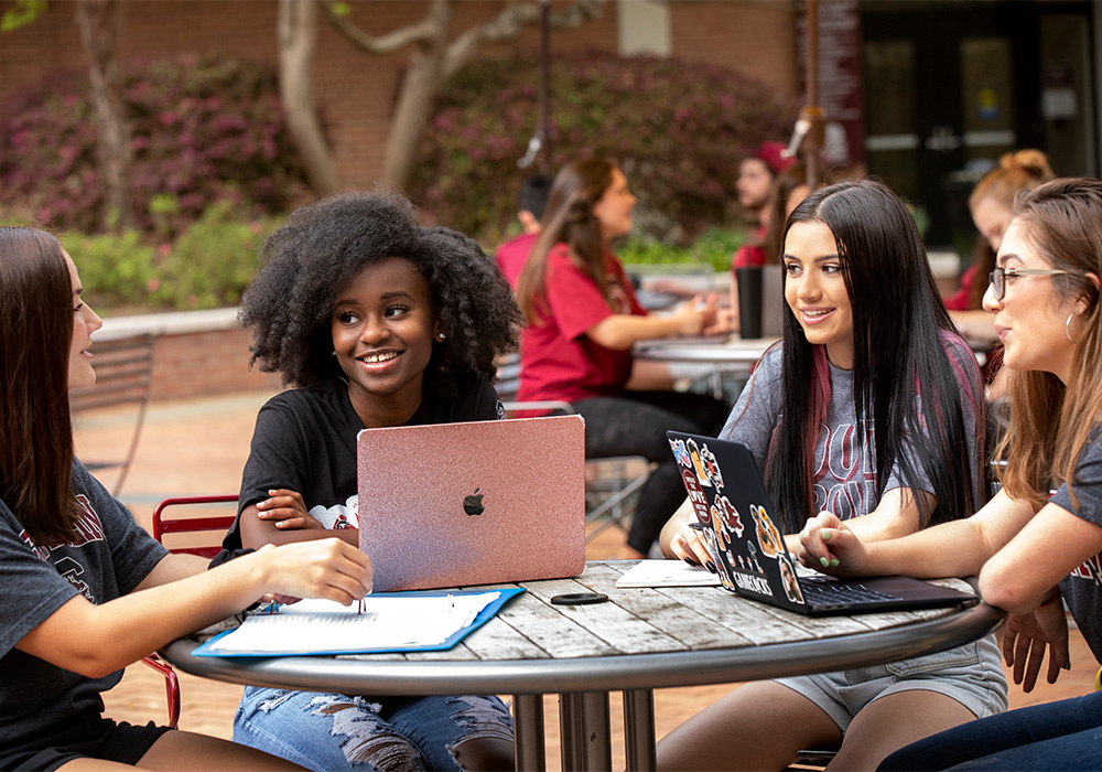 Group of students sitting at an outdoor table with laptops and notebooks.