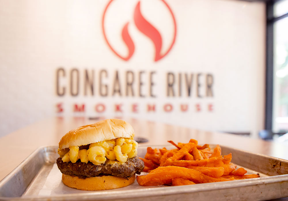 A tray with a mac and cheese burger with a side of sweet potato fries in front of the Congaree River Smokehouse sign. 