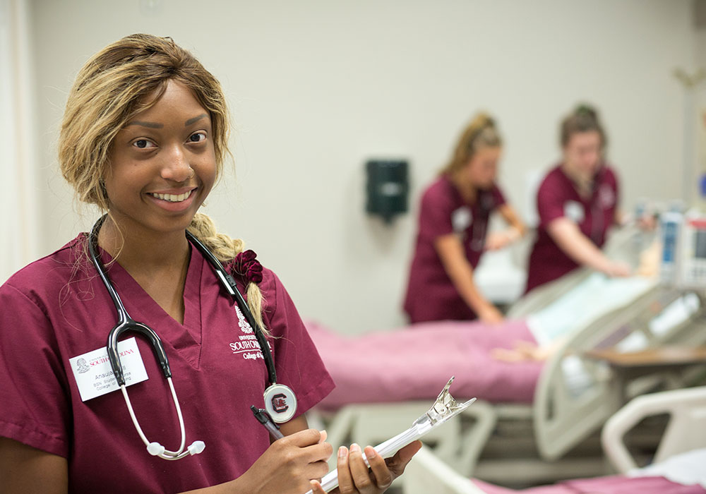 a smiling nursing student in a garnet uniform wearing a stethoscope, holding. a clipboard