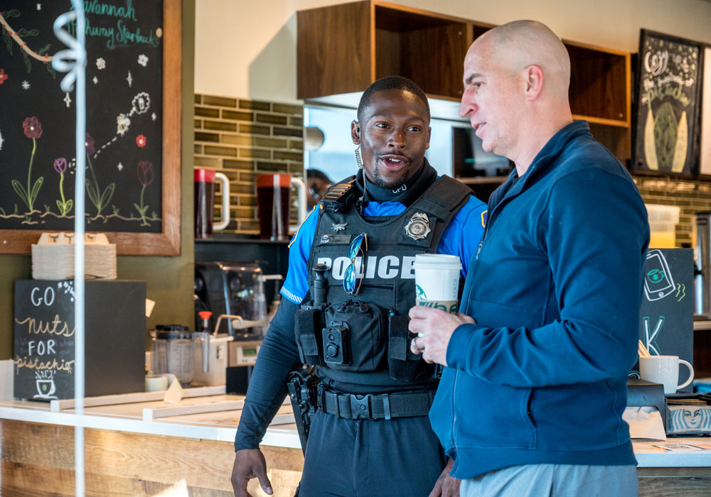 A police officer in uniform and tactical vest chats with a man holding a coffee cup inside a cafe.
