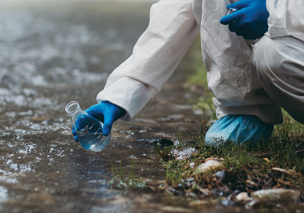 A person wearing porotectivce clothing gathers a water sample from the edge of a stream.