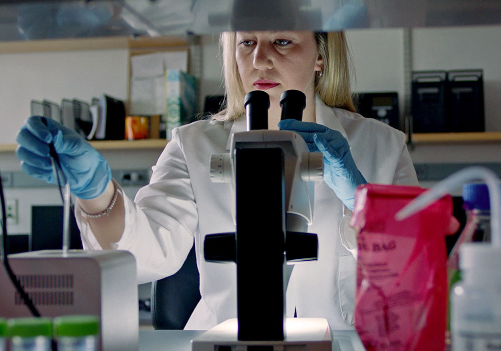 Video: Remarkable Research at USC. Photo: Woman in lab coat and gloves in a laboratory holding a microscope.