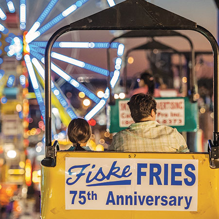 Two people riding a chairlift over the South Carolina State Fair in the evening surrounded by glowing lights from the other fair rides. 