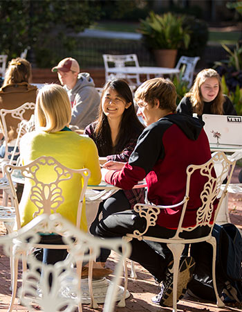 Clusters of students sit at various outdoor patio tables. 