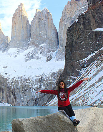 Student in a Gamecock shirt sitting on a bolder in front of a lake and a snowy mountain.