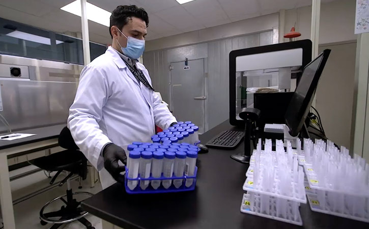 Student wearing a lab coat and safety equipment holds trays of test tubes in a lab setting. 